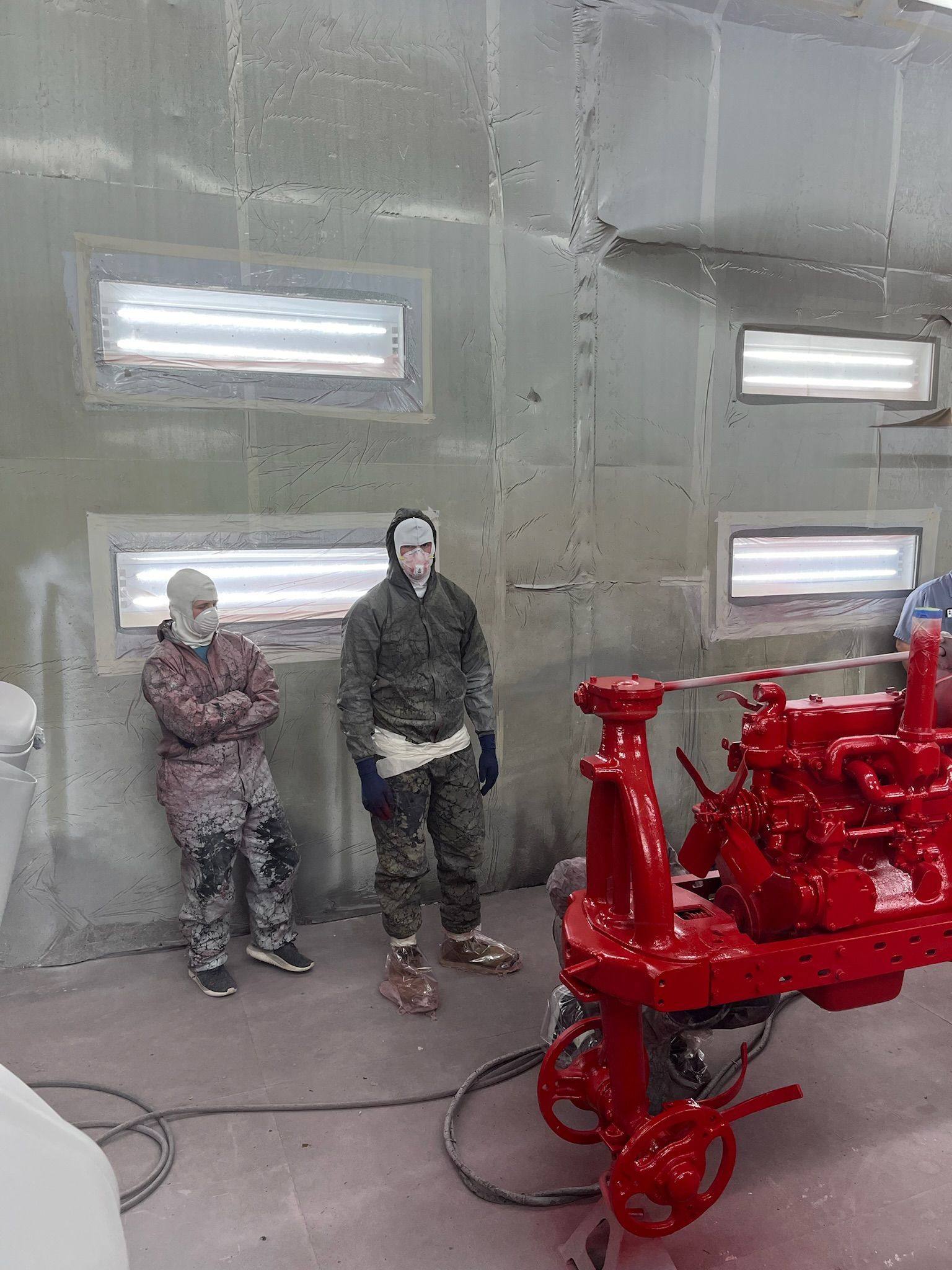 Two people stand near a red painted machine in a workshop. Fluorescent lights and a gray wall are visible.