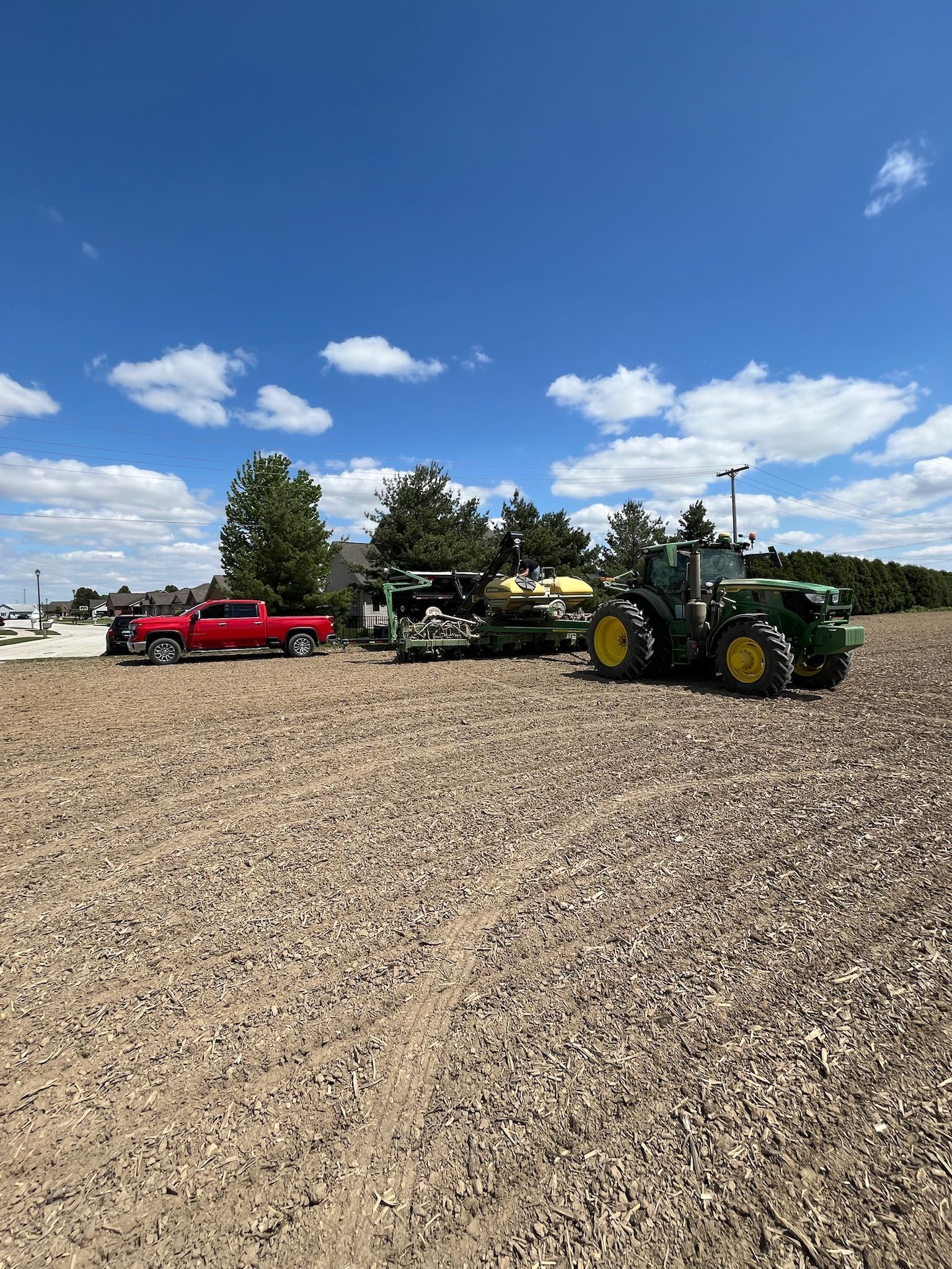 Red truck, trailer, and green John Deere tractor on a field under a blue sky.