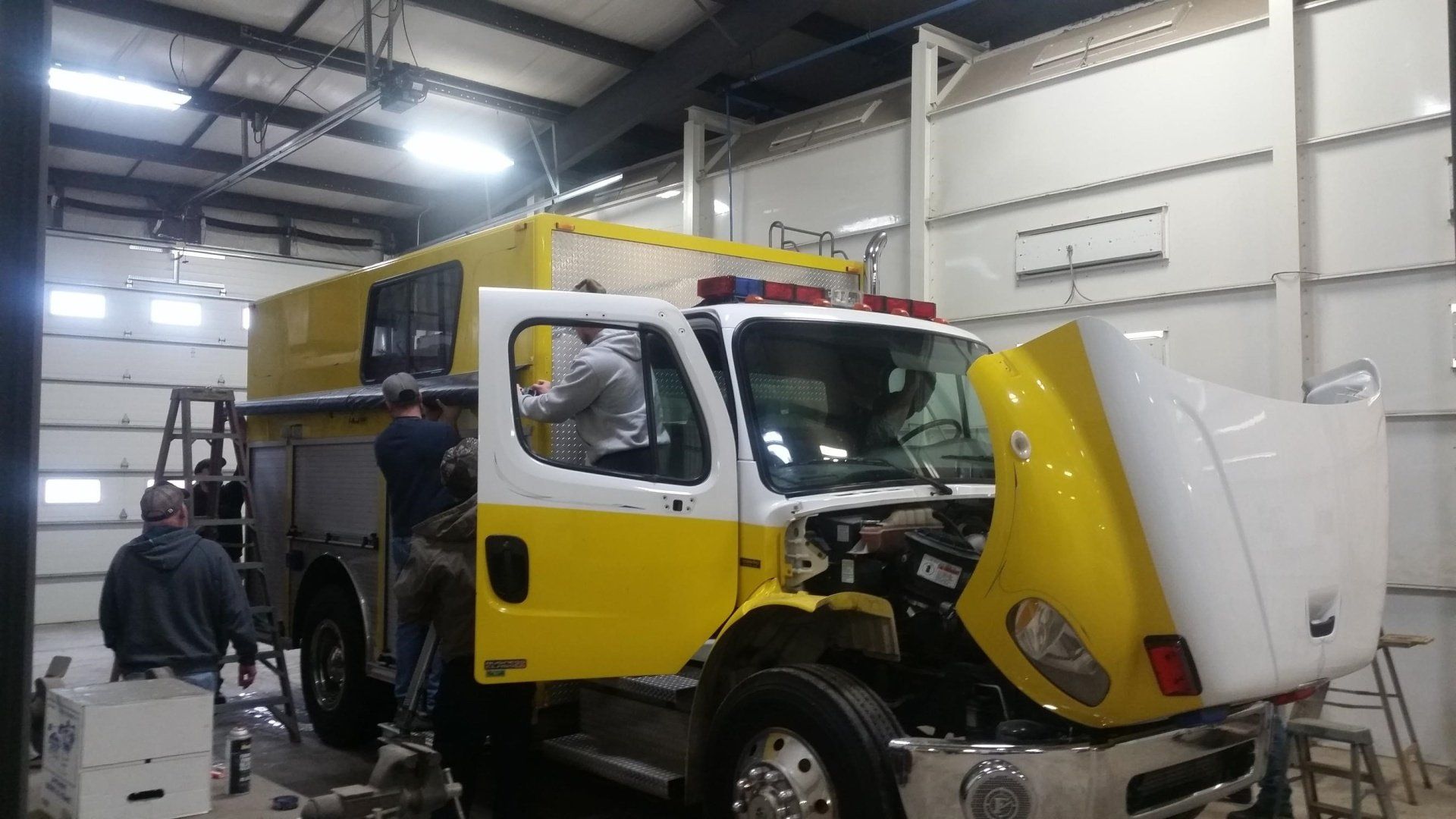 Fire truck being worked on in a garage; yellow and white paint scheme; workers visible.