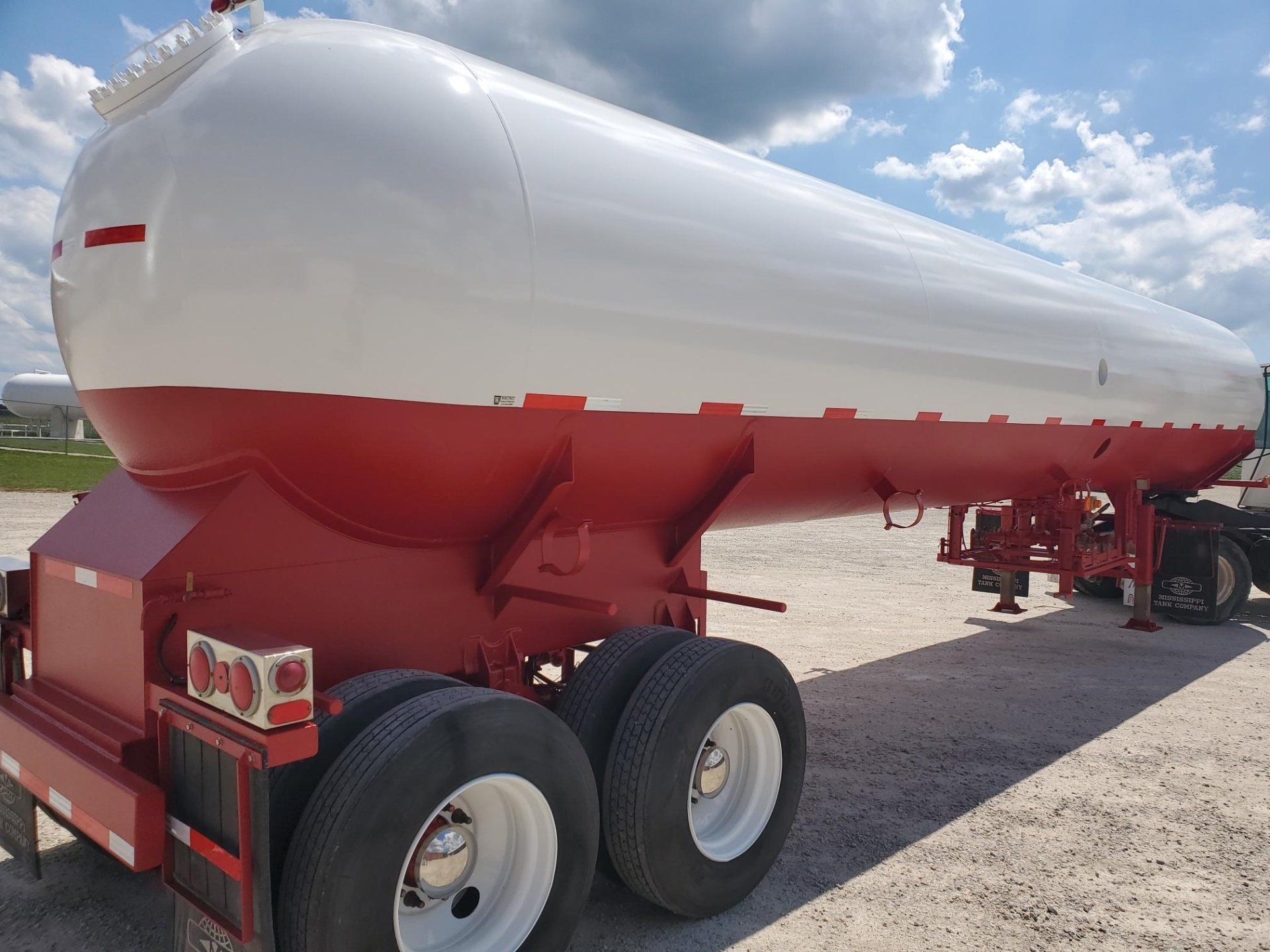 Red and white propane tanker trailer on a gravel lot under a partly cloudy sky.