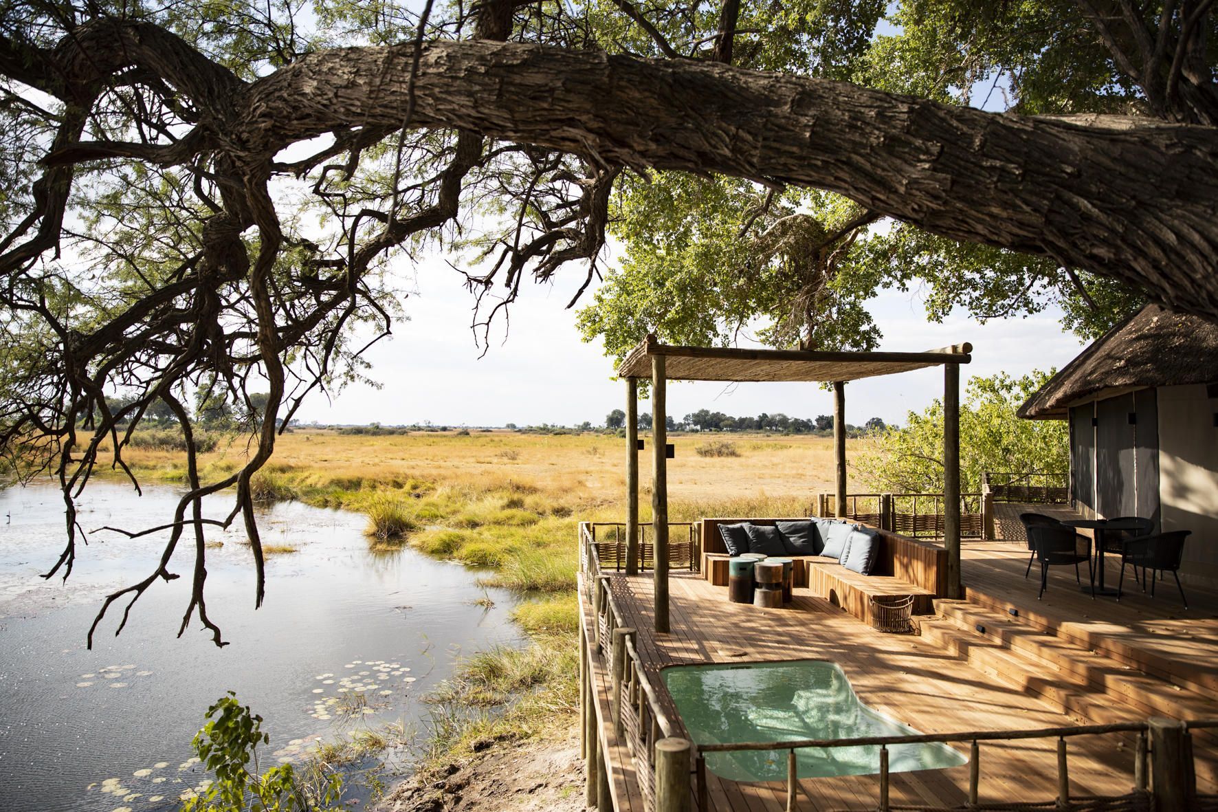 A scenic wooden deck with an outdoor pool overlooks a calm river and vast golden plains under a large, leafy tree.