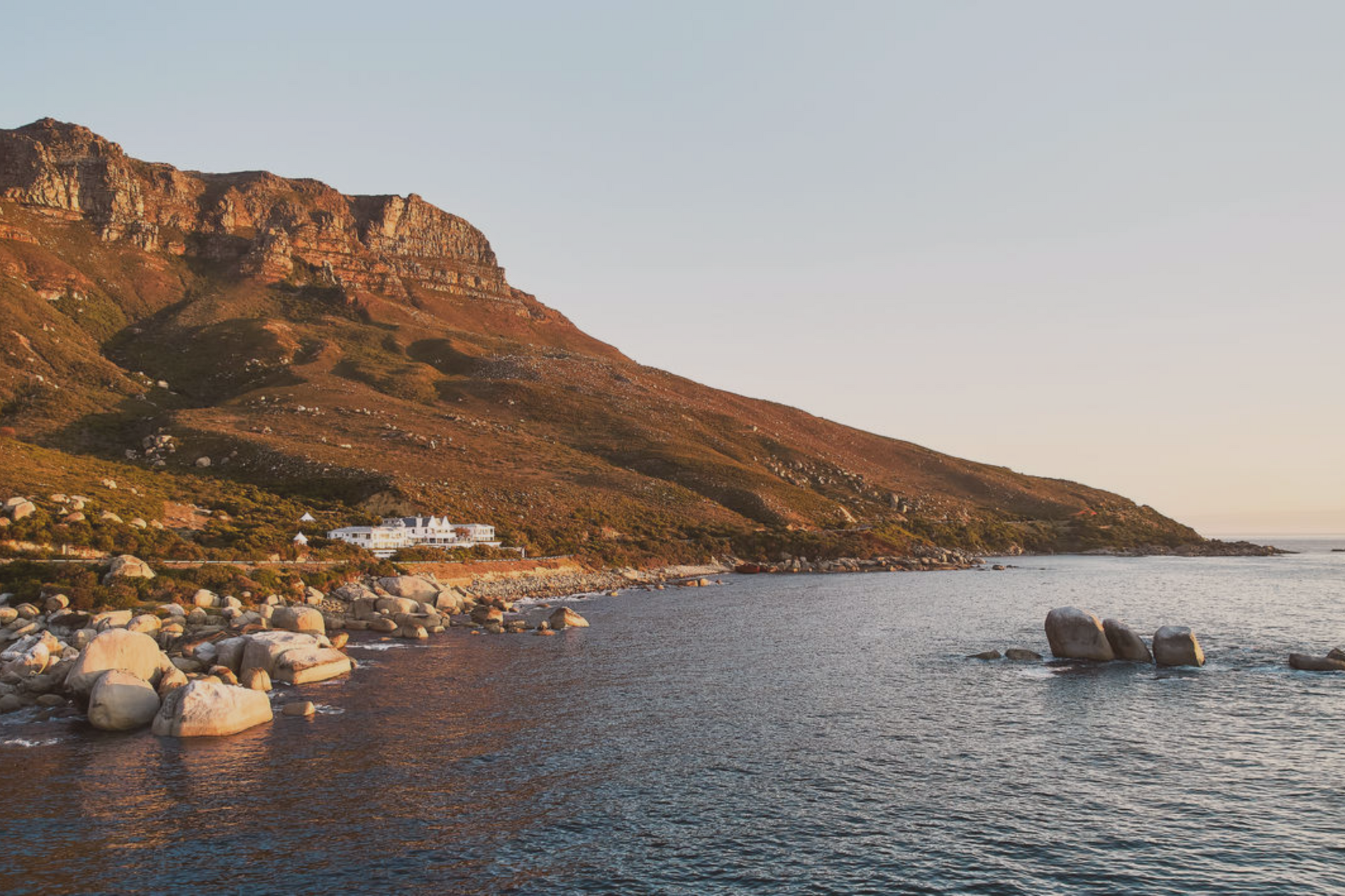 A scenic coastal view featuring a rocky shoreline, blue water, and a large, sunlit mountain slope.