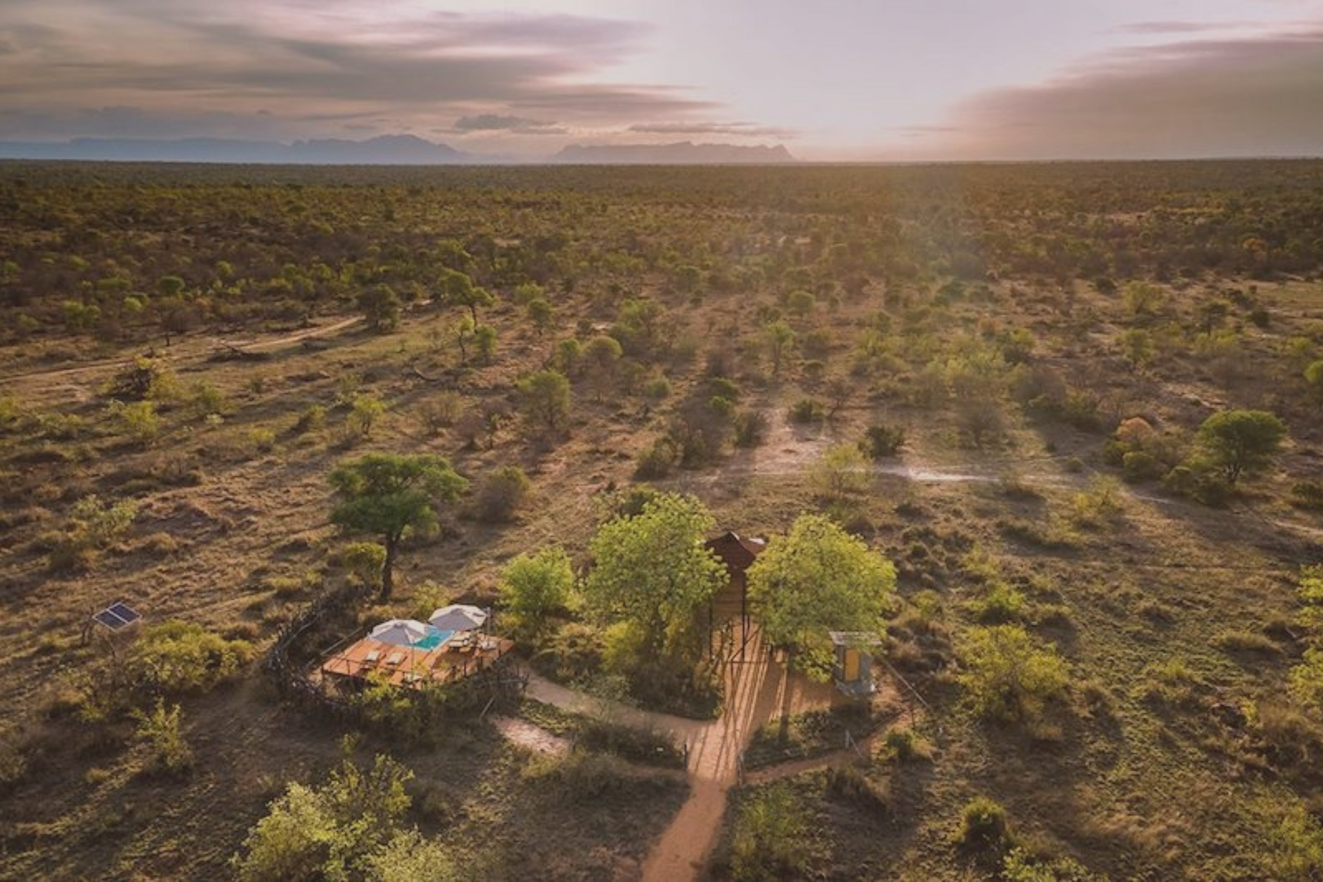 Aerial view of a remote safari lodge with a swimming pool nestled in a lush, sunlit savanna landscape at sunset.