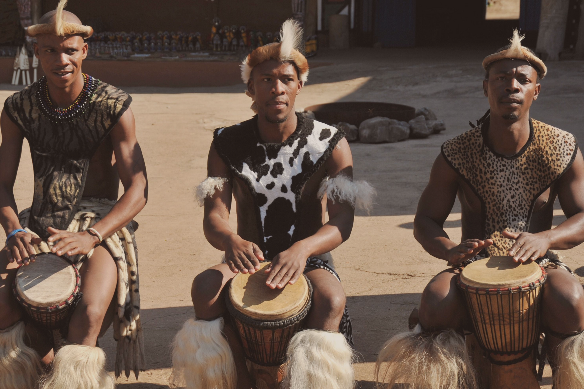 Three musicians in traditional African attire sit on the ground, each playing a hand drum in an outdoor setting.