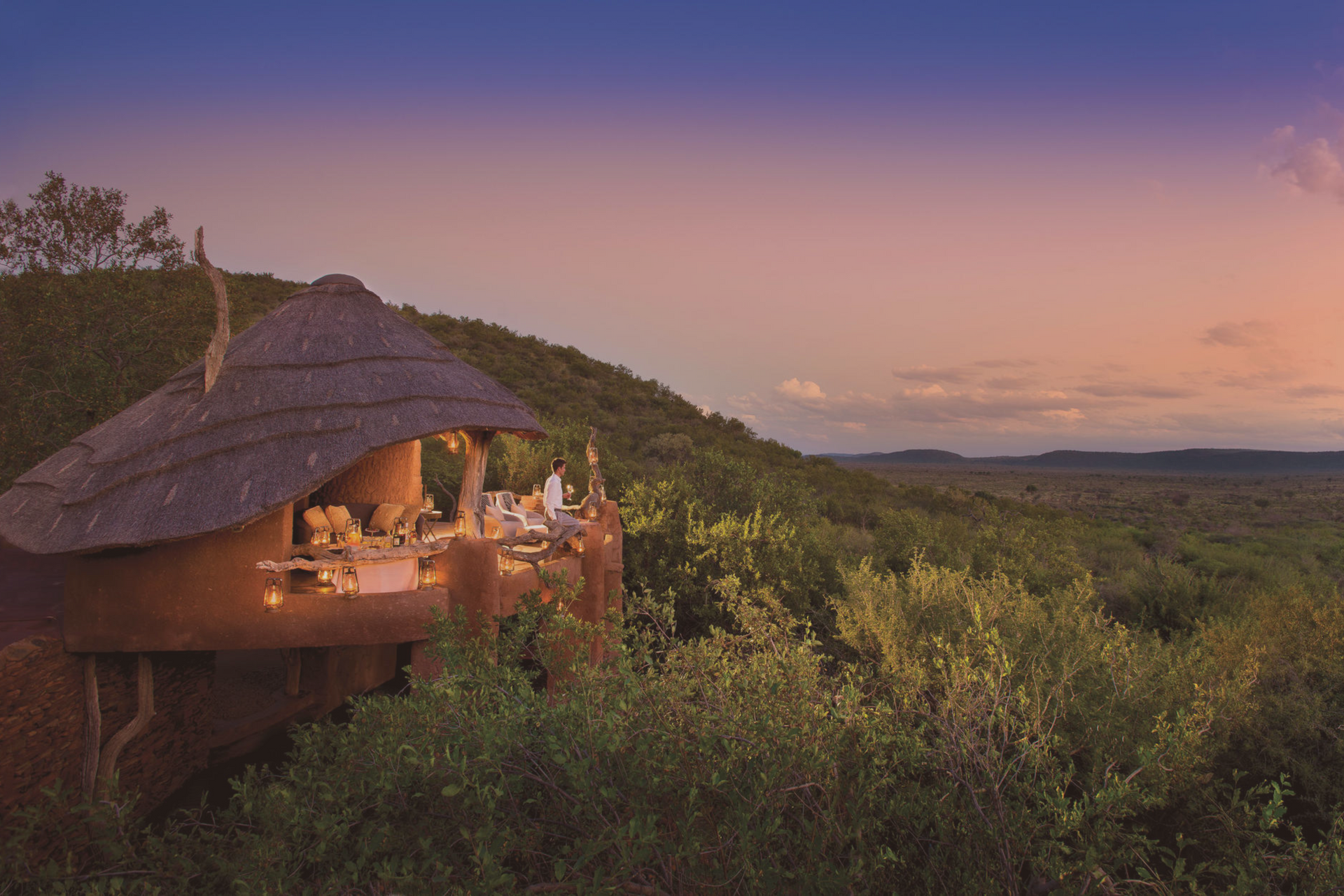 A sunset view of a thatched-roof treehouse balcony overlooking a vast, lush forest landscape.