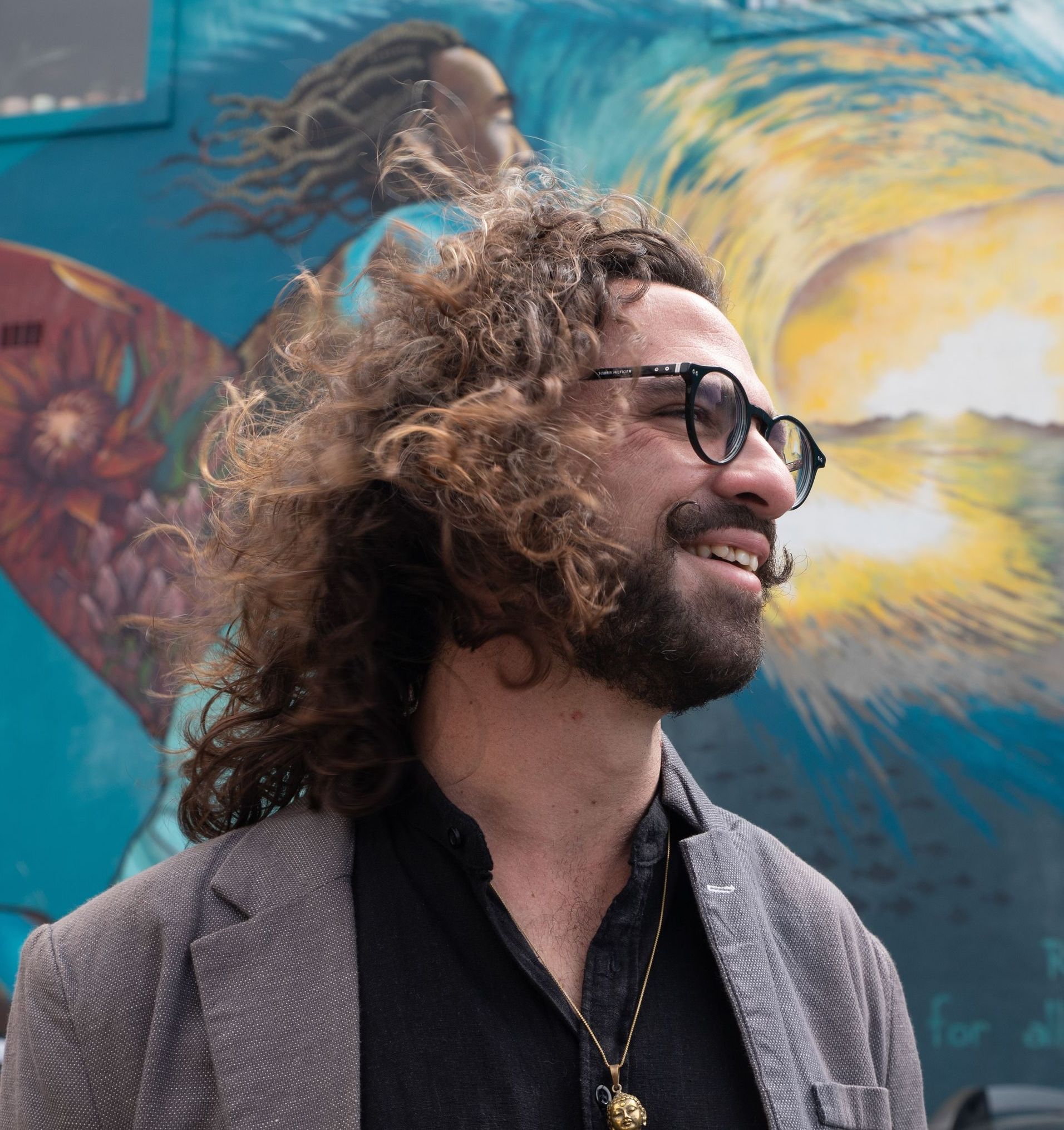 A man with curly hair and glasses is smiling in front of a mural