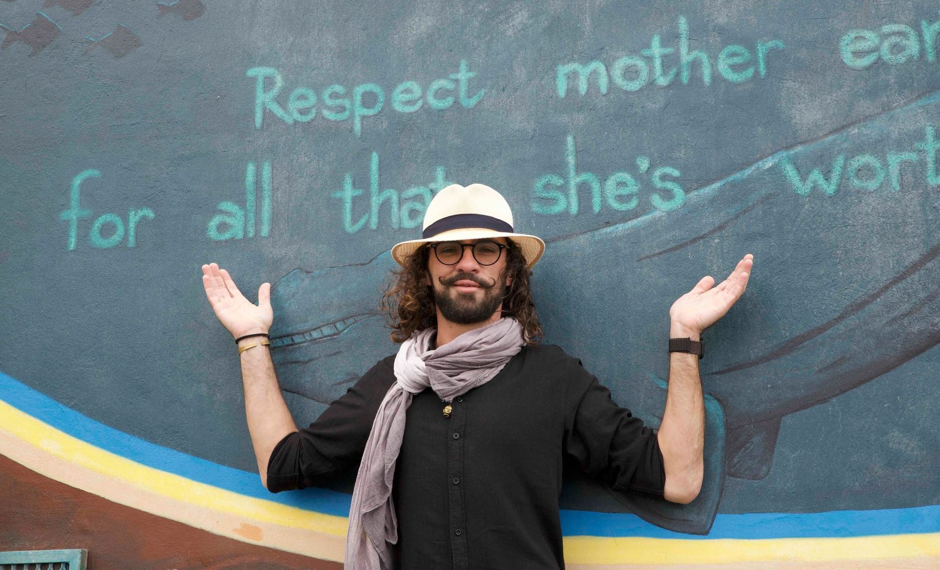 Conserrvation travel agent standing before a colorful mural with raised arms and handwritten environmental message.