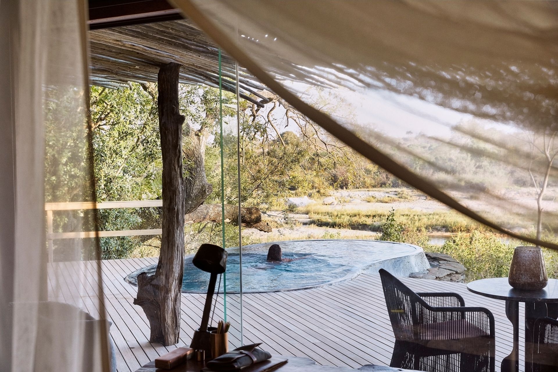 A person relaxing in an outdoor pool on a wooden deck overlooking a savanna landscape, viewed through a draped tent.