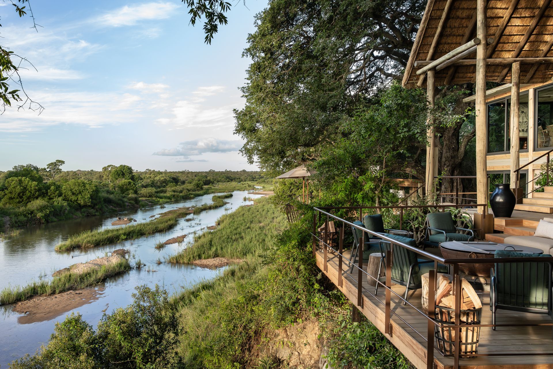 An elevated wooden deck with patio furniture overlooks a river winding through a lush, green African savanna landscape.