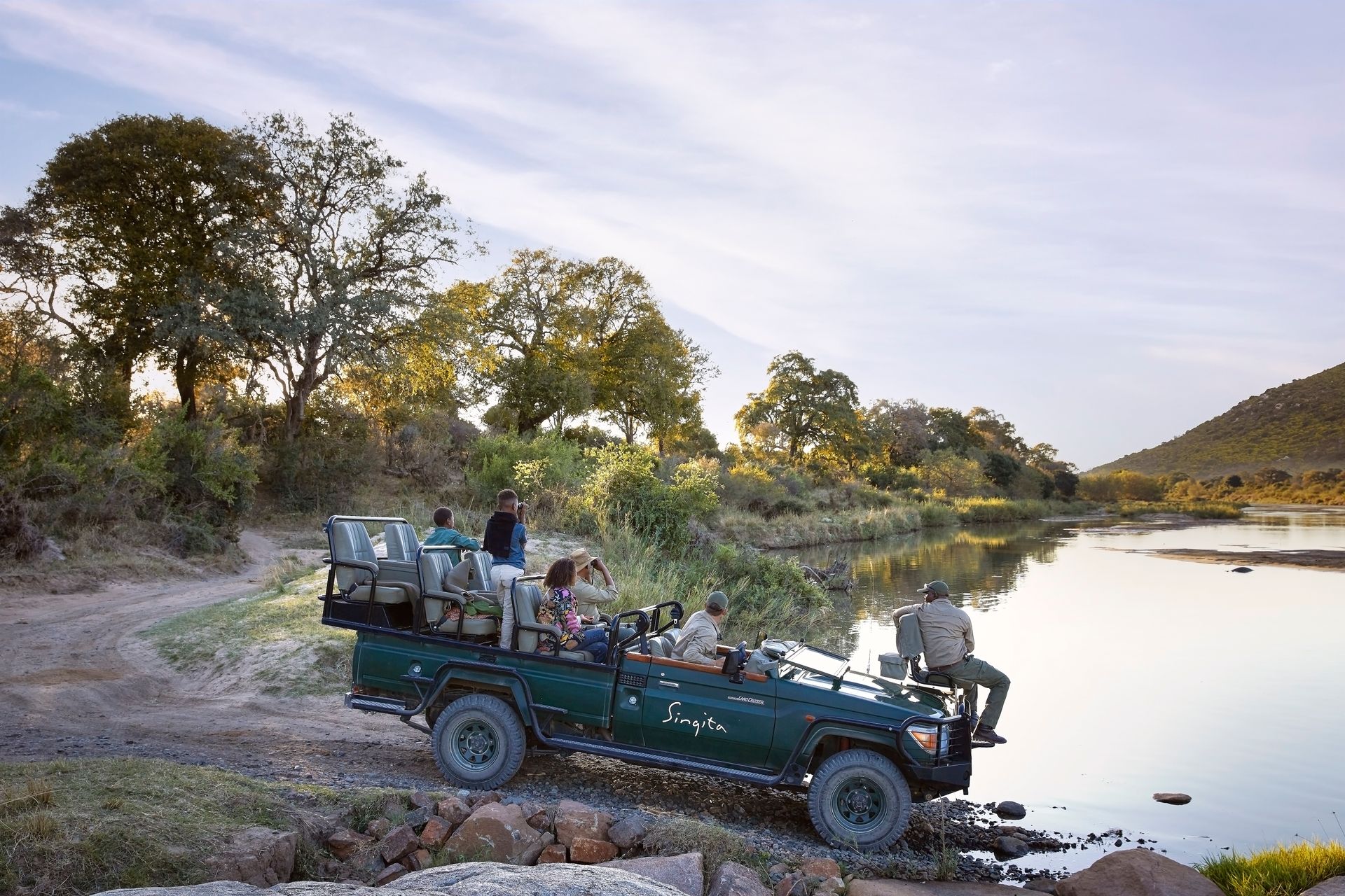 A group of people in an open-topped safari vehicle parked by a river in a wooded, hilly landscape.