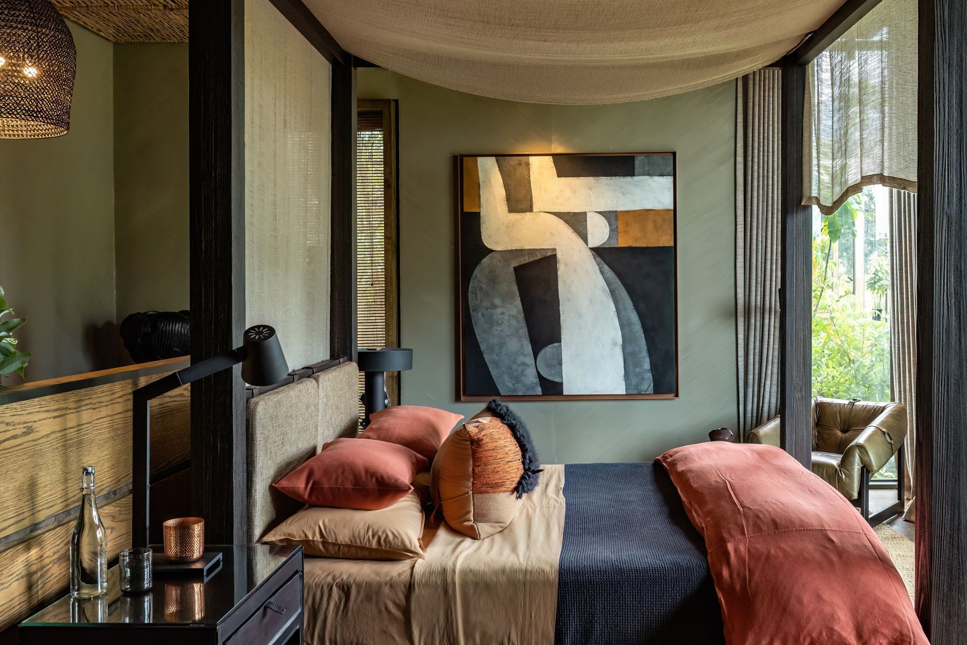 A modern bedroom with earthy tones, a canopy bed, abstract wall art, and a large window looking out onto greenery.