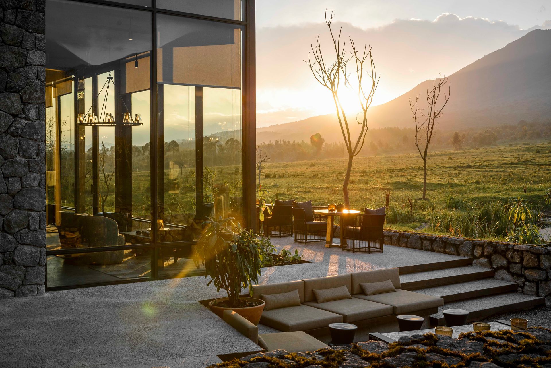 Modern stone-walled building with large glass windows overlooking a grassy field and mountain at sunset.