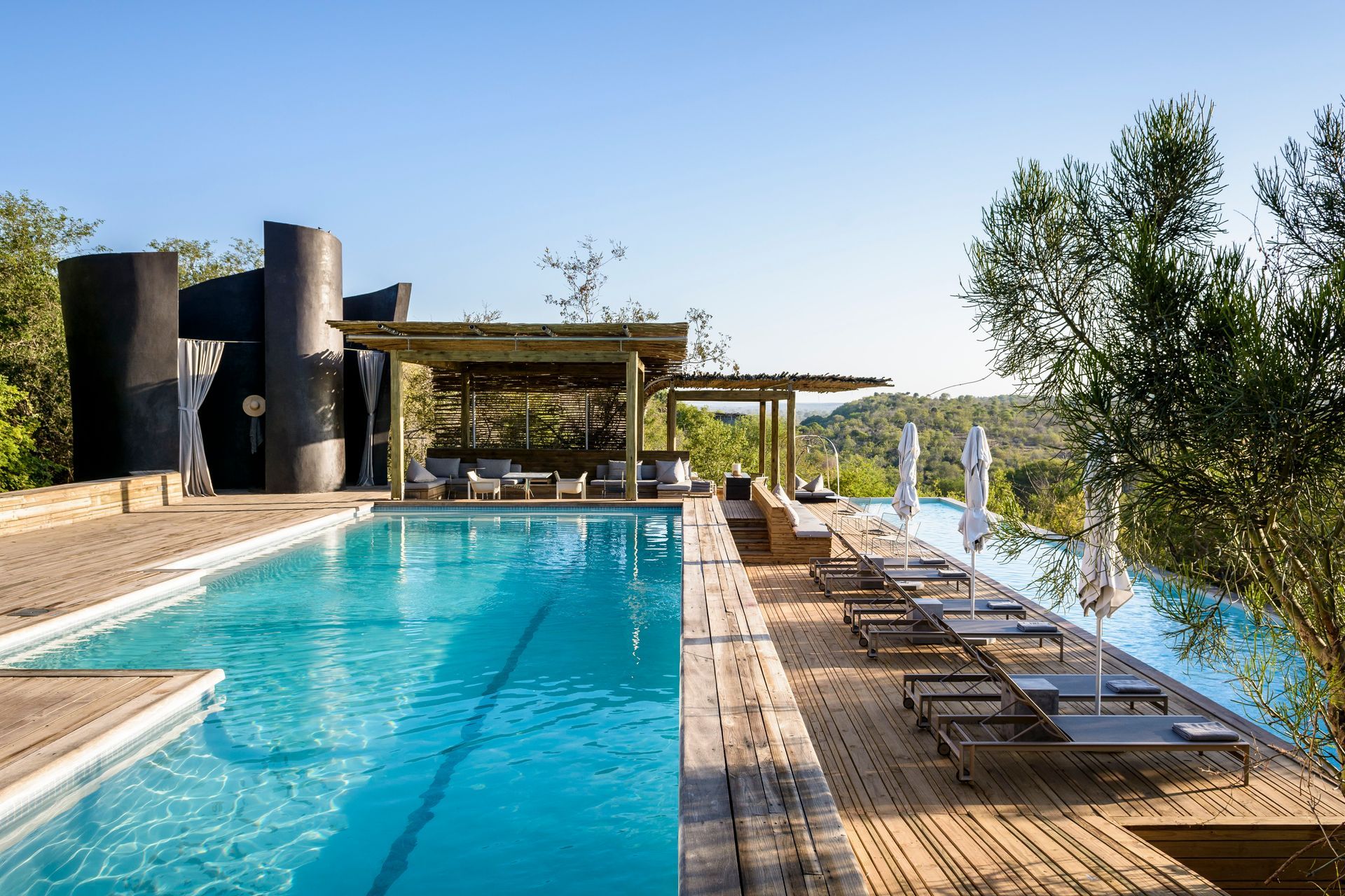 A long, rectangular infinity swimming pool lined with lounge chairs, overlooking a hilly landscape on a sunny day.