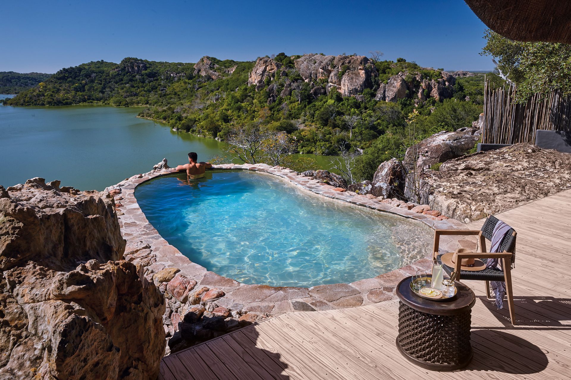 A person relaxing in a stone-edged infinity pool overlooking a scenic lake, rocky hillside, and lush greenery.