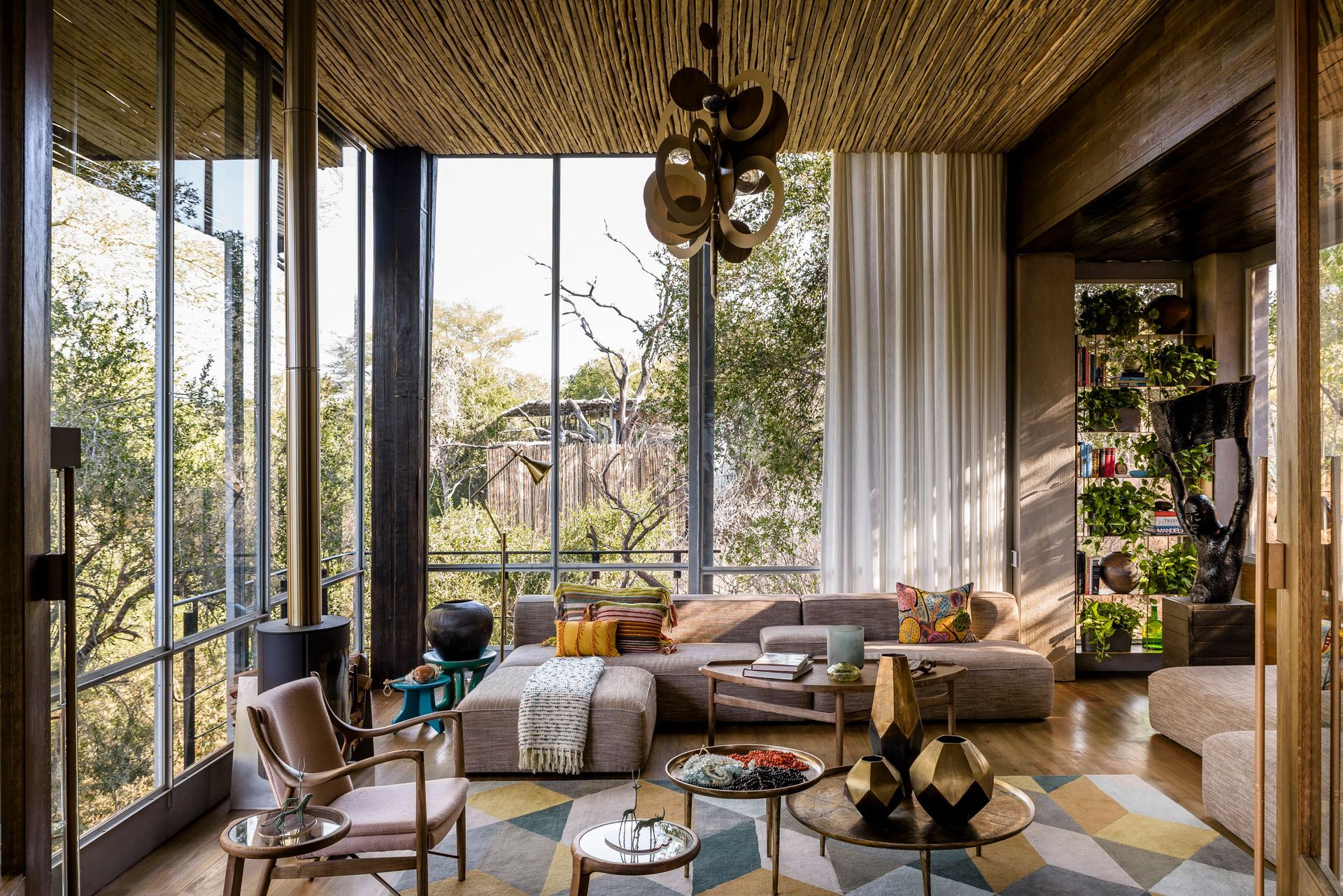 A modern living room with large glass walls, a textured ceiling, a sectional sofa, and floor-to-ceiling indoor plants.