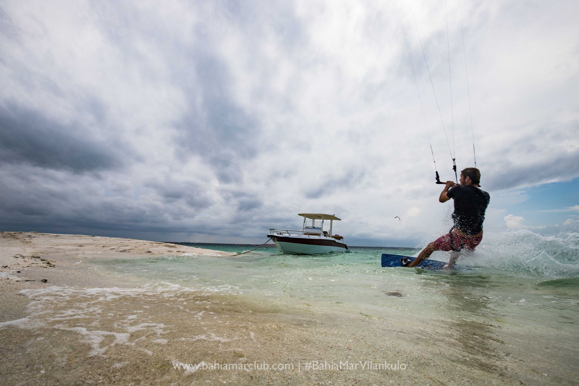 A man is flying a kite in the ocean with a boat in the background.