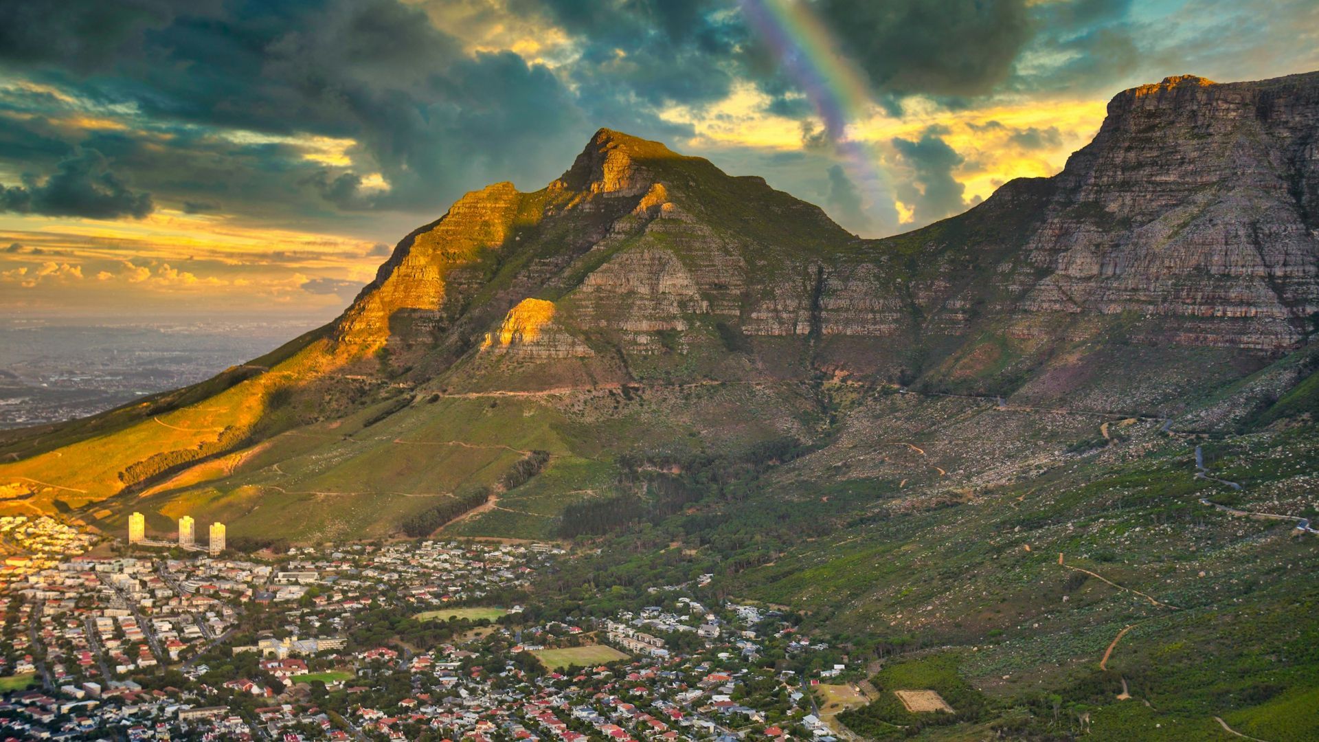 A city is surrounded by mountains and a rainbow is visible in the sky.