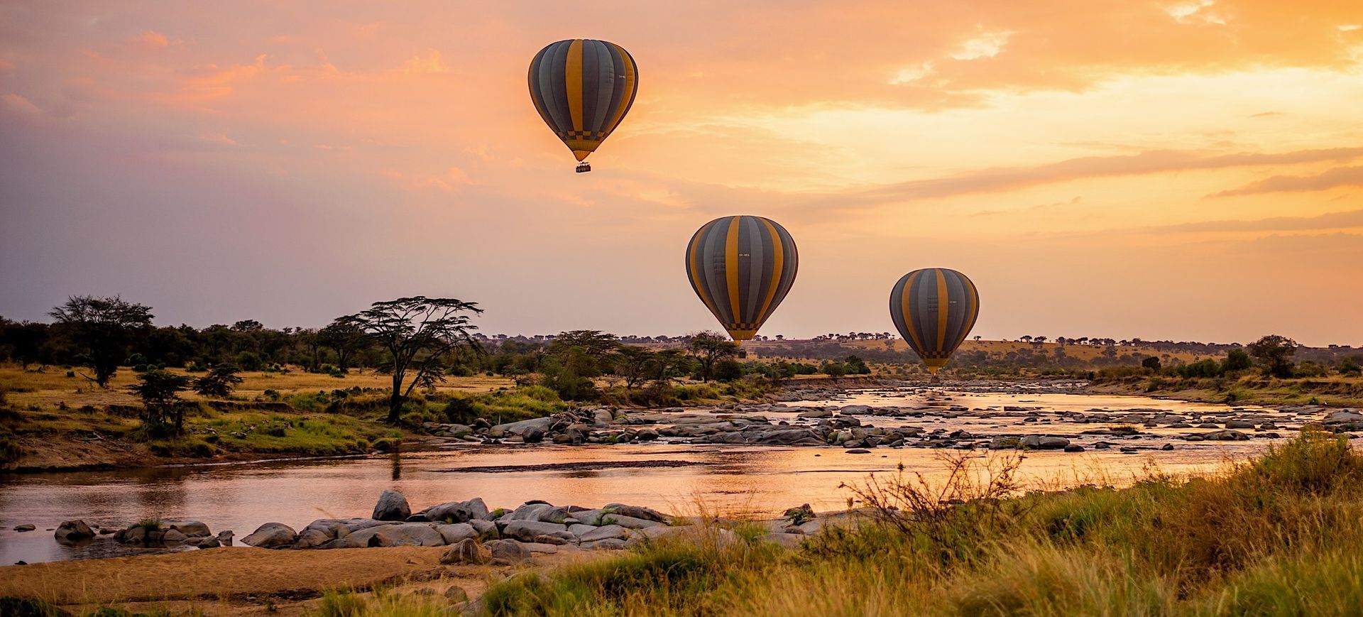 Three hot air balloons are flying over a river at sunset.