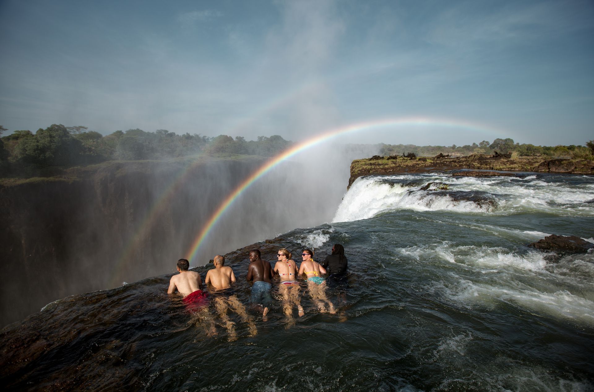 A group of people are sitting in the water near a waterfall with a rainbow in the background.