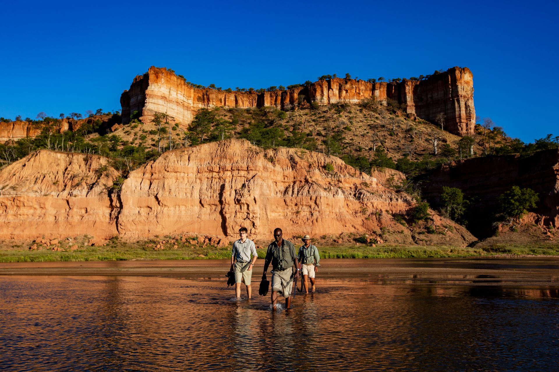 A group of people are walking through a body of water with a mountain in the background.
