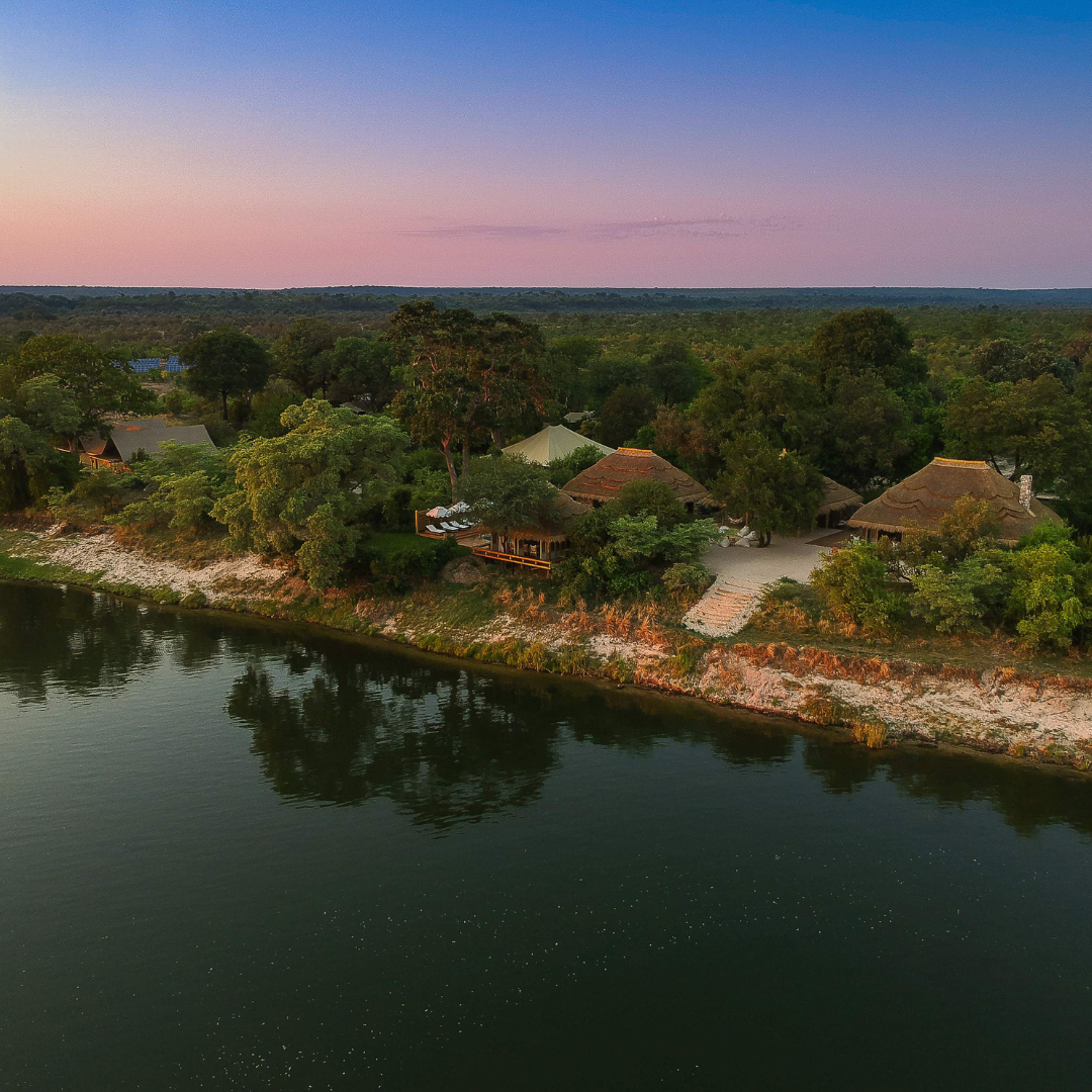 Aerial view of thatched-roof lodges nestled along a riverbank at dusk under a soft pink and blue sky.