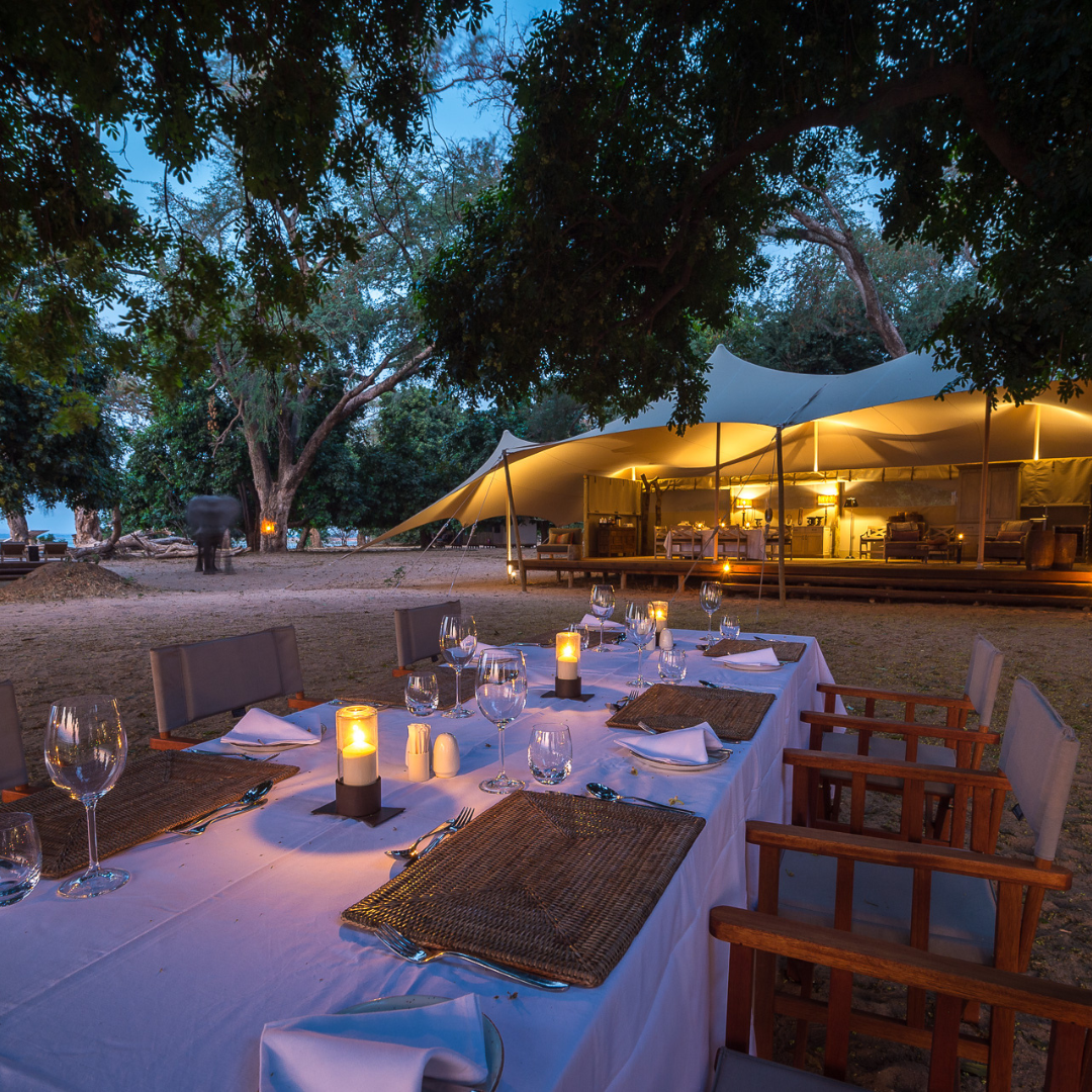 A long dining table set for an outdoor dinner at dusk, with a lit canvas safari tent visible in the background.