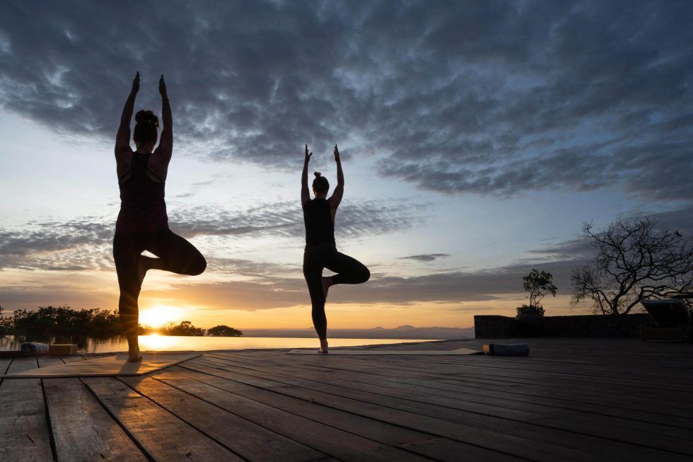 Two women are doing yoga on a wooden deck at sunset.