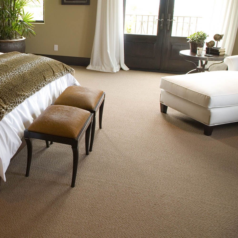 Bedroom with tan carpet, a bed, two ottomans, and a white upholstered chair.