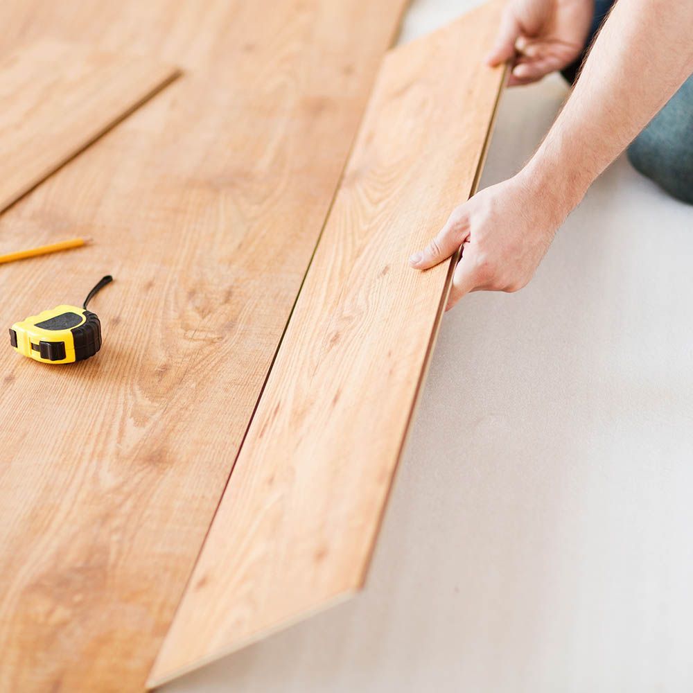 Person installing wood flooring, holding a plank next to laid planks. Yellow measuring tape and pencil visible.
