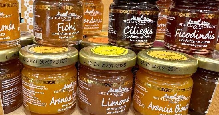 Glass jars of fruit preserves with gold lids, labeled Fichi, Ciliegia, and Arancia, stacked on display.