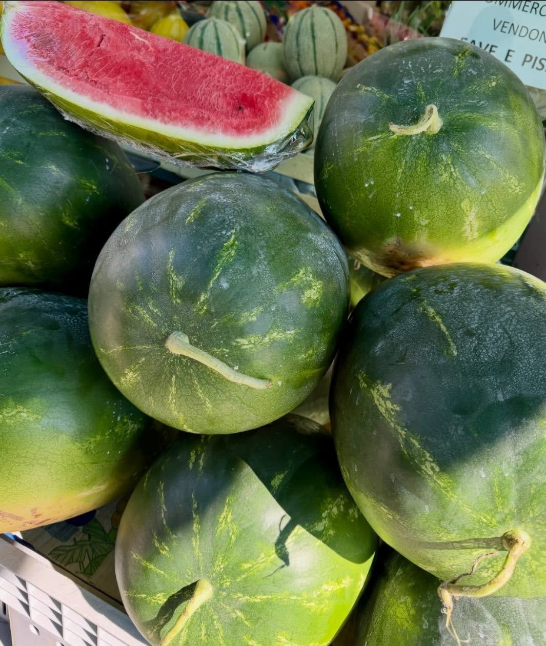 Watermelons piled at a market, with one cut open showing bright red flesh.