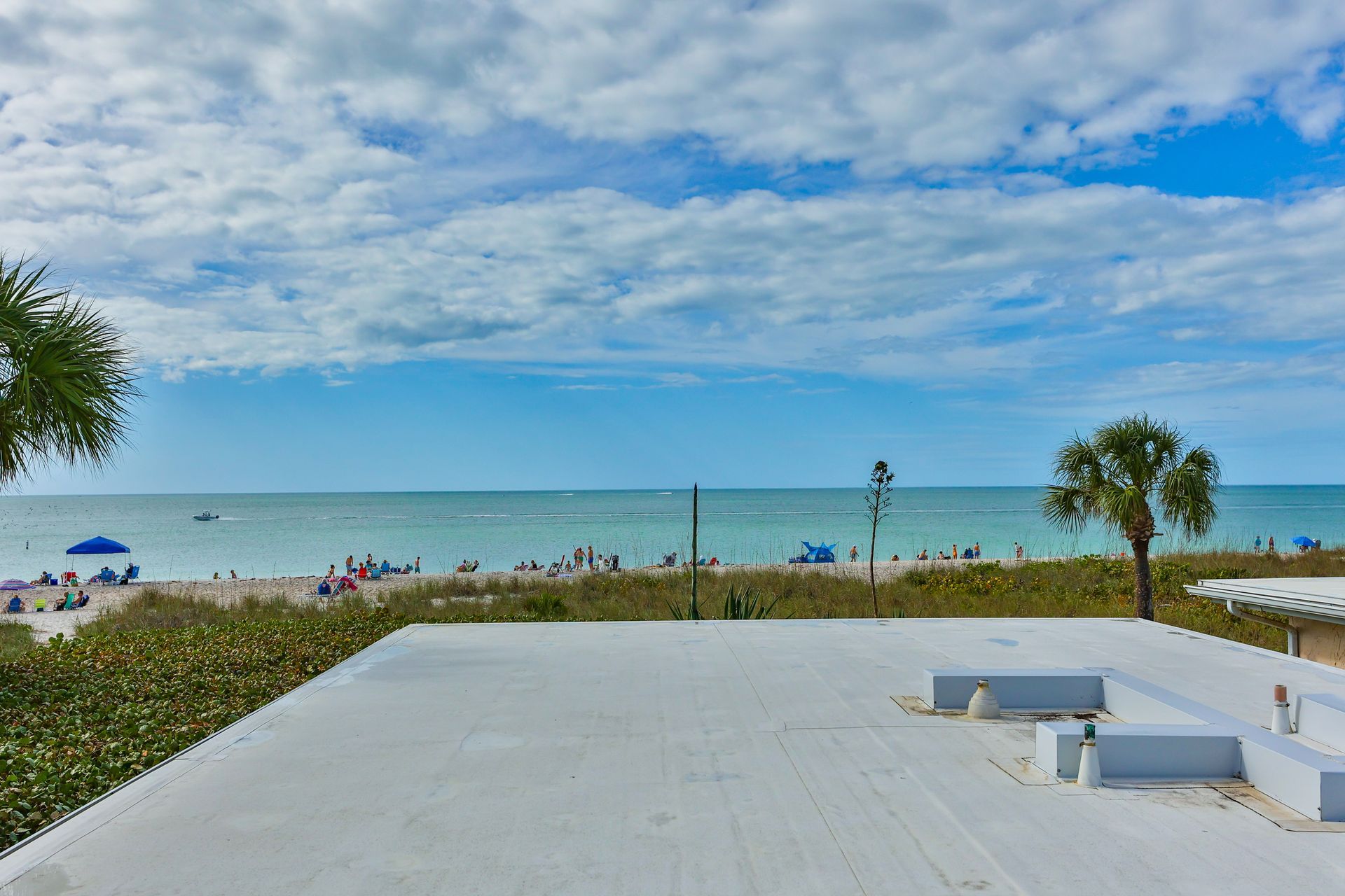 Beach view from a rooftop: blue sky, white clouds, sandy beach, ocean, people, palm trees, and a flat roof.