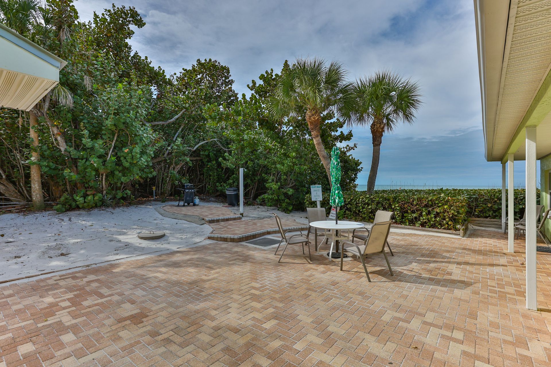 Patio with table, chairs, beach, and foliage. Cloudy sky.