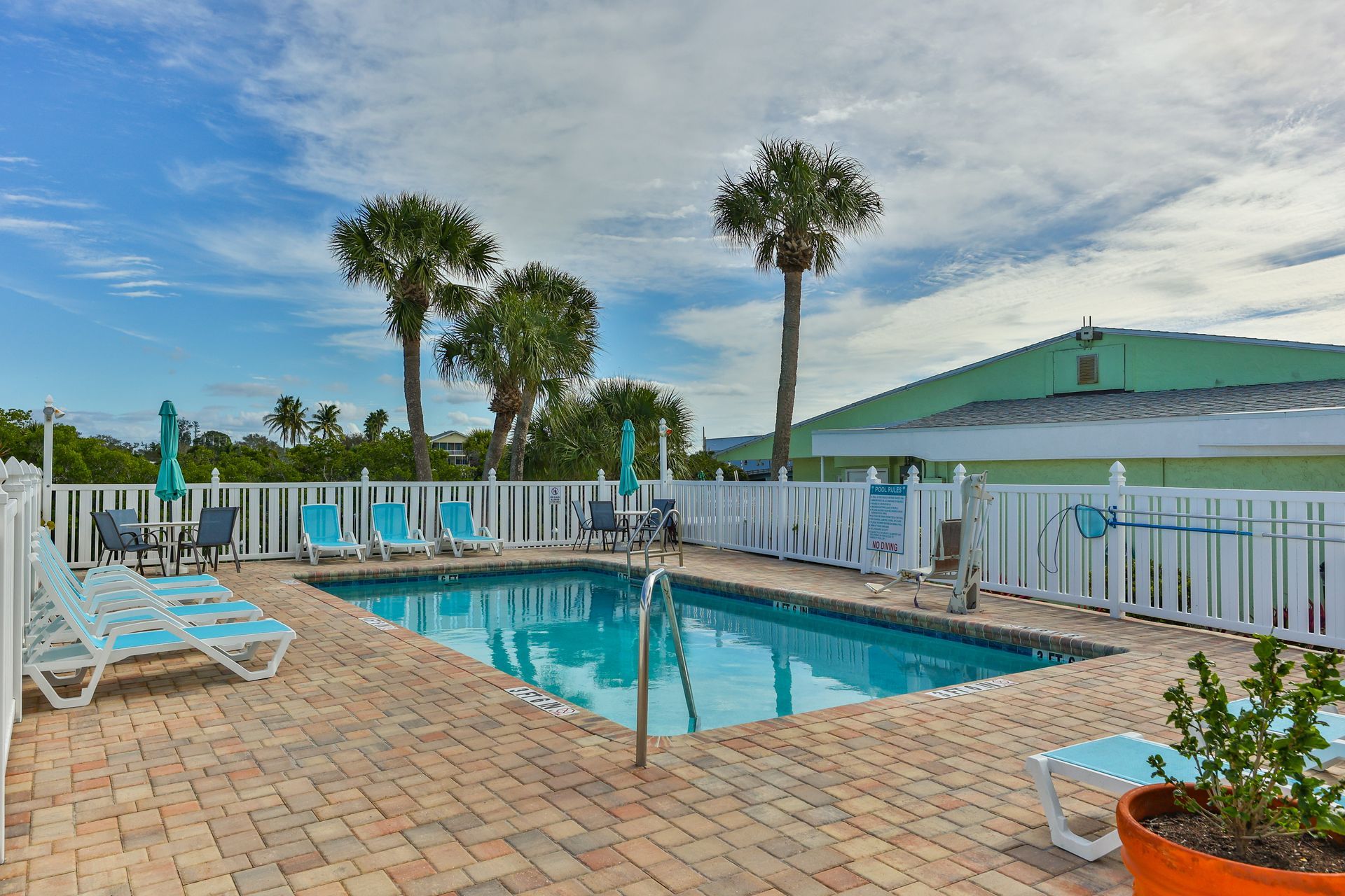Pool with lounge chairs, palm trees, and a white fence against a cloudy blue sky.