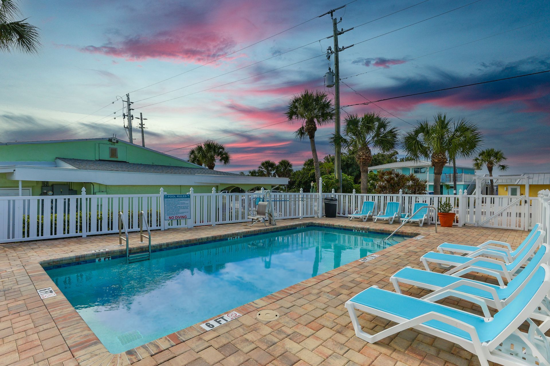 Poolside scene: blue pool, lounge chairs, white fence, green building, sunset sky.