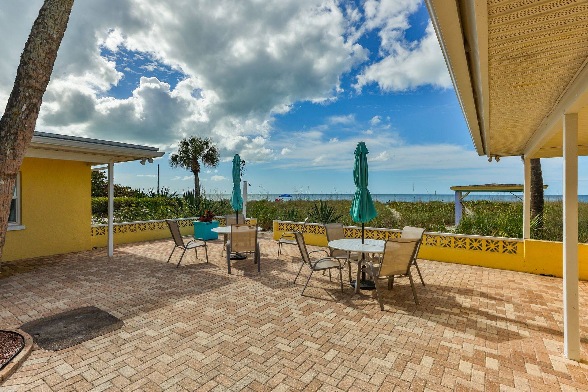 Patio with tables, chairs, and umbrellas overlooking a beach with blue sky and clouds.