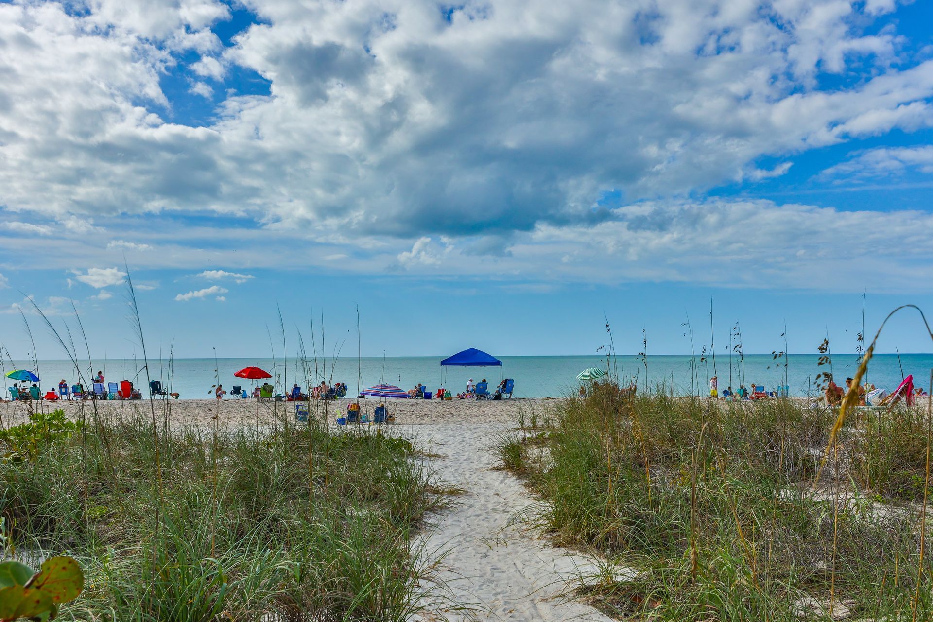 Sandy path leads to a beach with people, umbrellas, and the ocean under a cloudy blue sky.