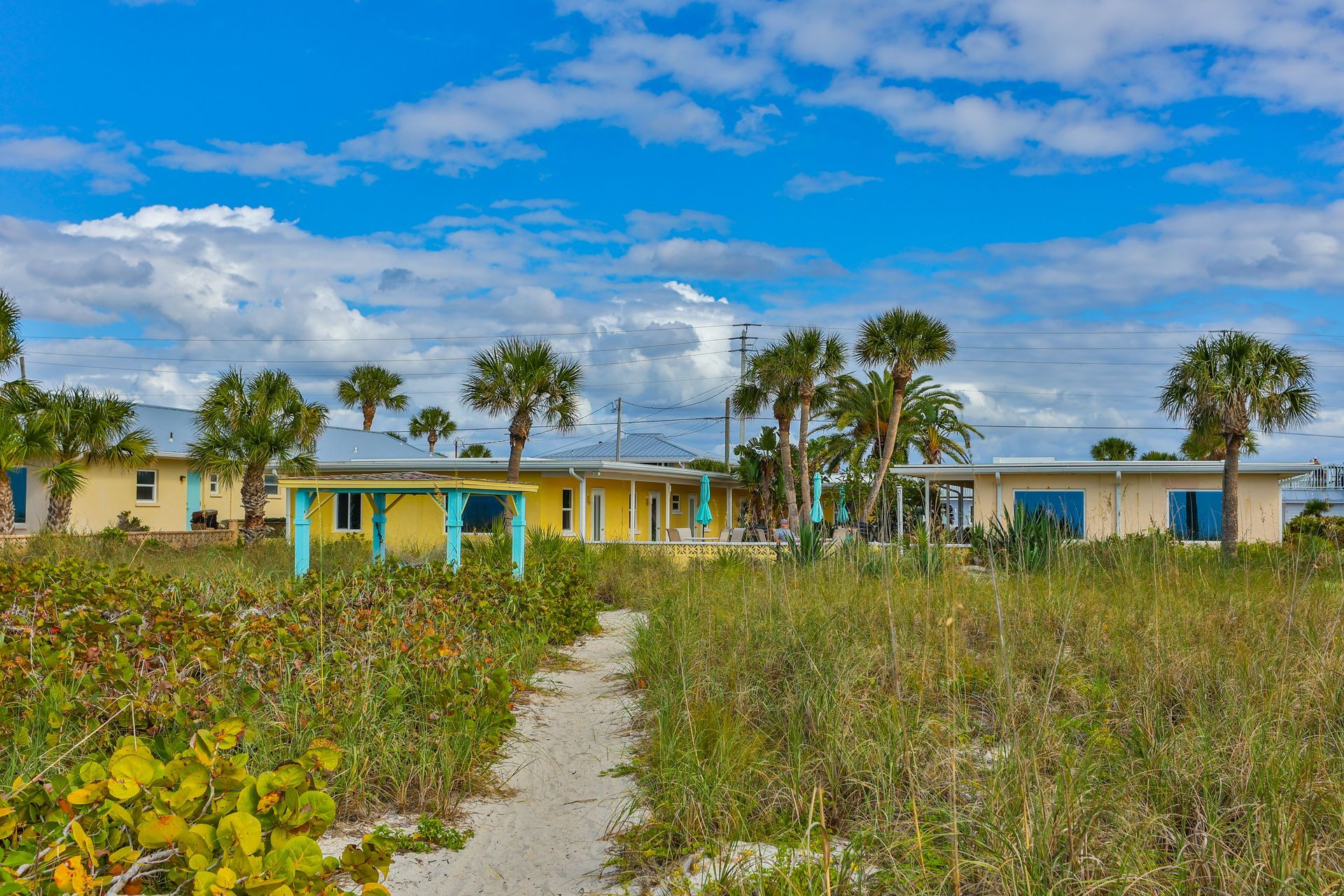 Sandy path leads to beachside houses under a bright blue sky with palm trees.