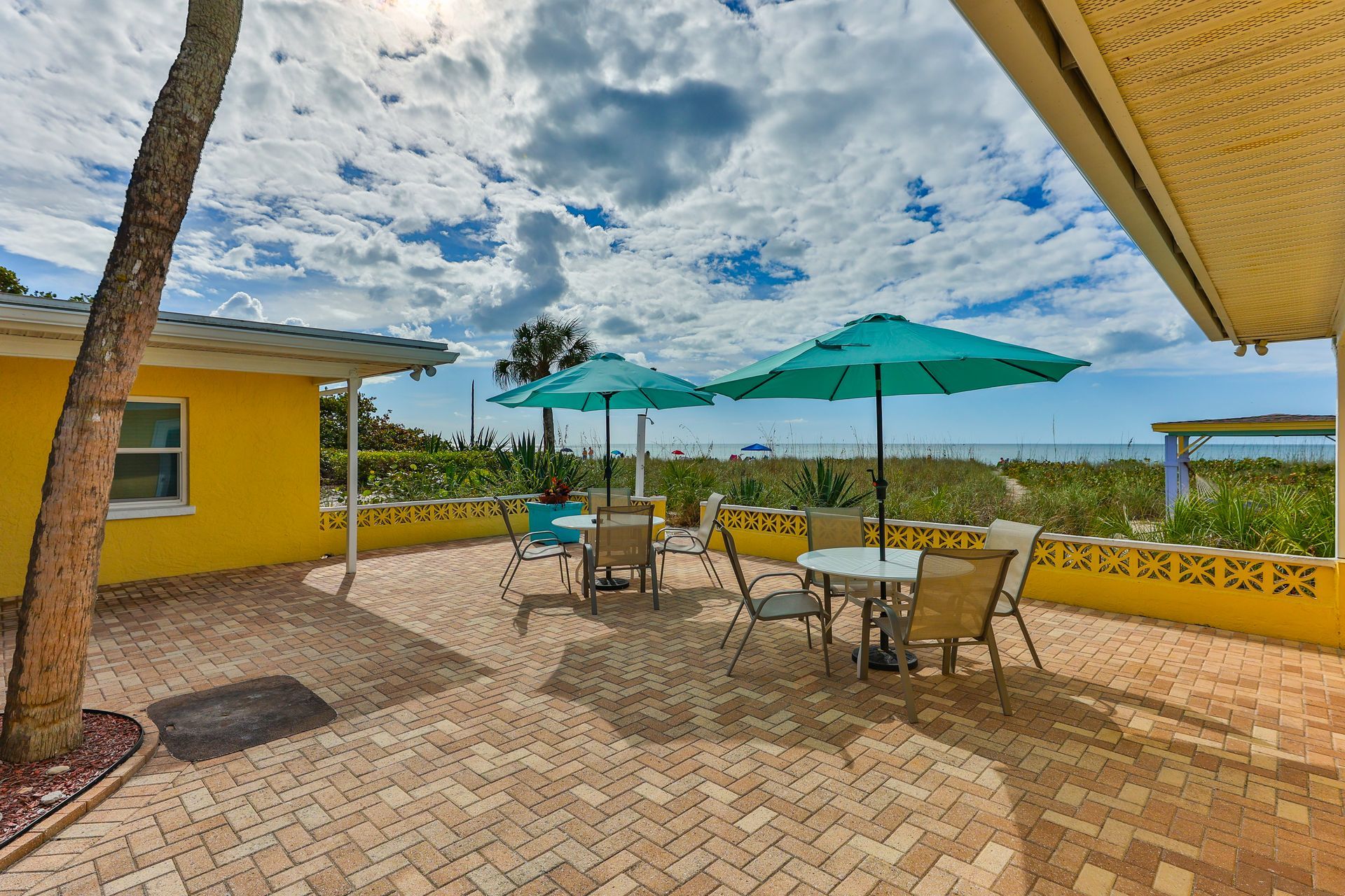 Patio with tables, chairs, teal umbrellas, and ocean view, next to a yellow building.