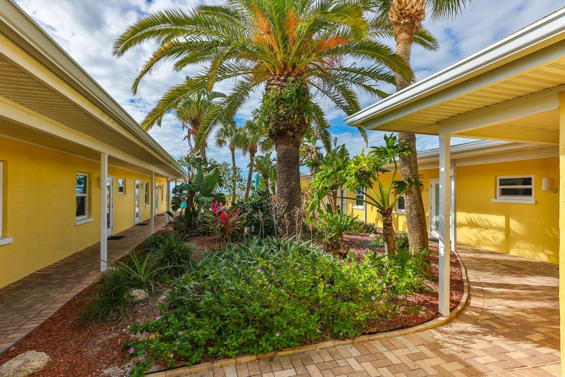 Yellow buildings with walkway, lush tropical plants, and palm trees under a bright blue sky.