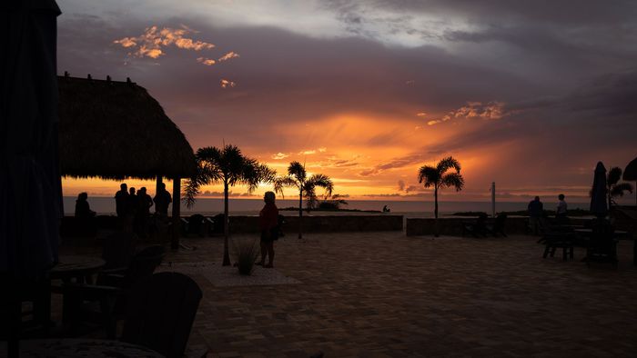 Sunset over a beach with palm trees, silhouetted people, and a thatched hut on the left