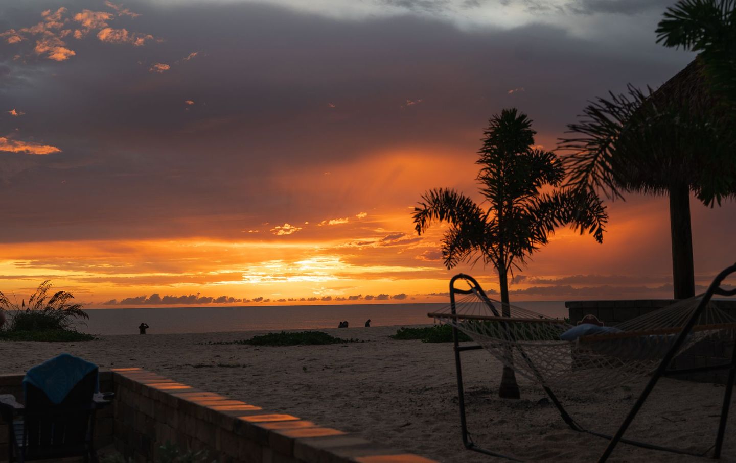 Sunset over a sandy beach with palm trees and lounge chairs silhouetted against orange clouds