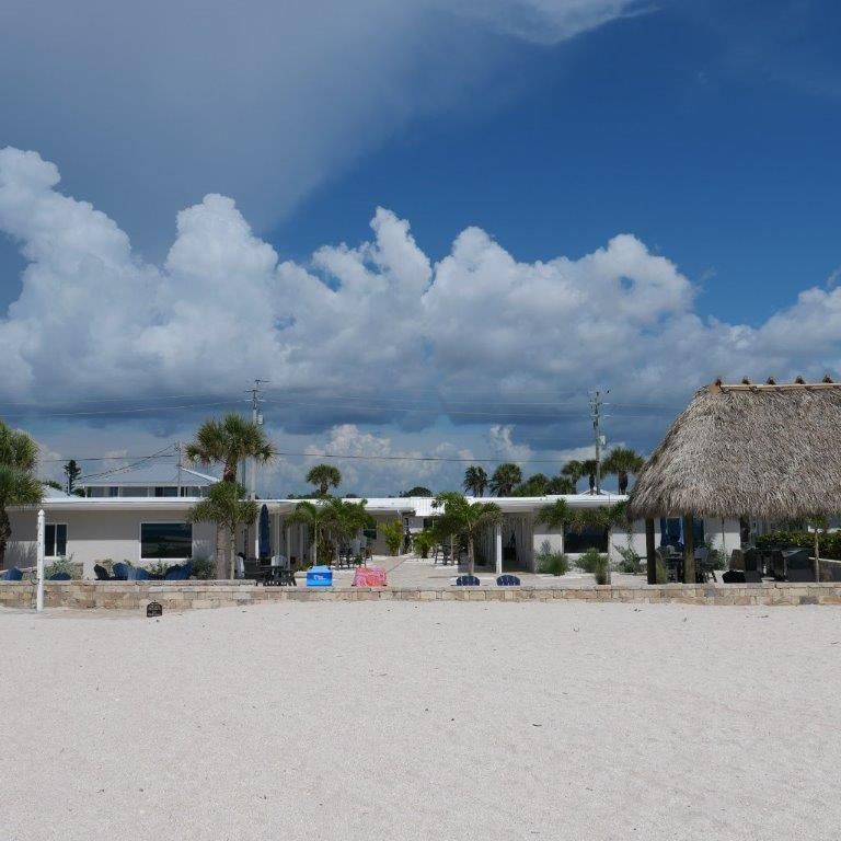 Sandy beach with white resort buildings, palm trees, colorful chairs, and a large cloudy blue sky