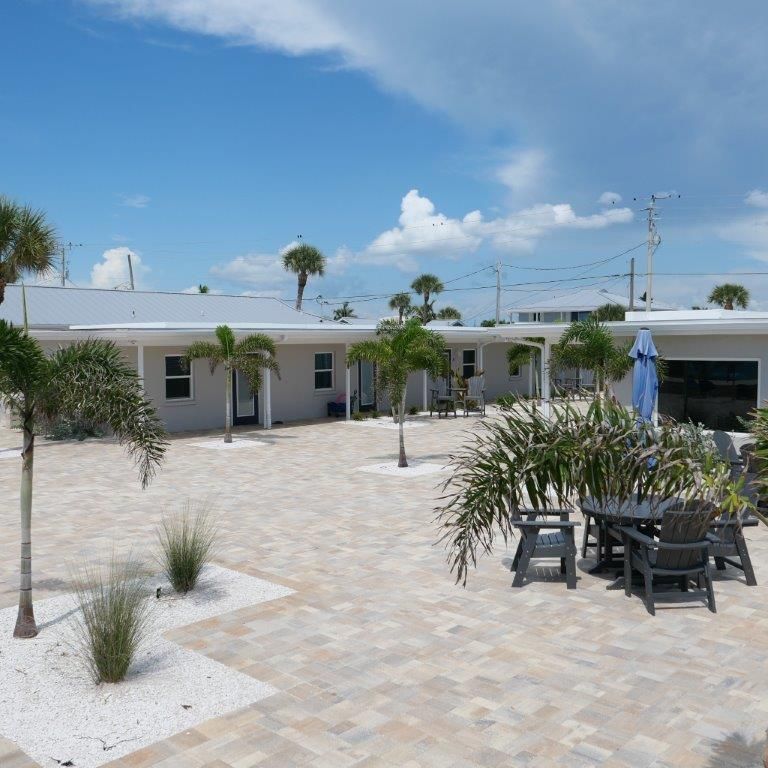 Sunny courtyard with white buildings, palm trees, and patio tables under a blue sky