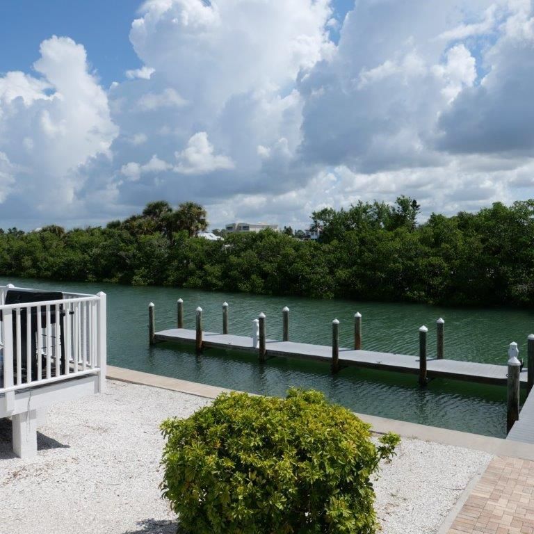 White dock with posts beside green water under a cloudy sky, viewed from a patio with a shrub.
