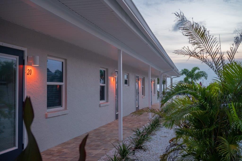 White motel corridor at dusk with lit wall lamps, windows, and palm trees along a gravel walkway