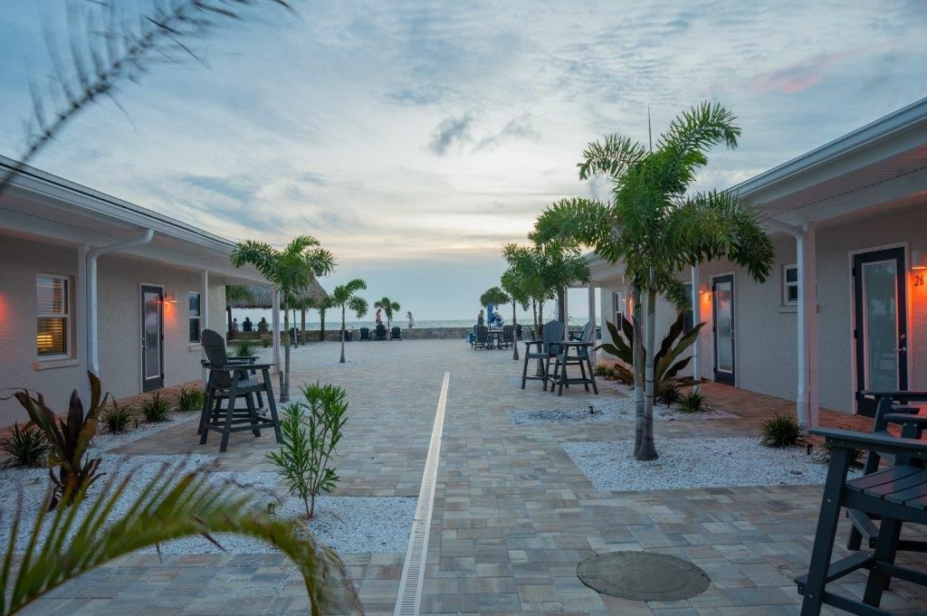 Courtyard walkway between white buildings at dusk, with palm trees, tables, and ocean view ahead