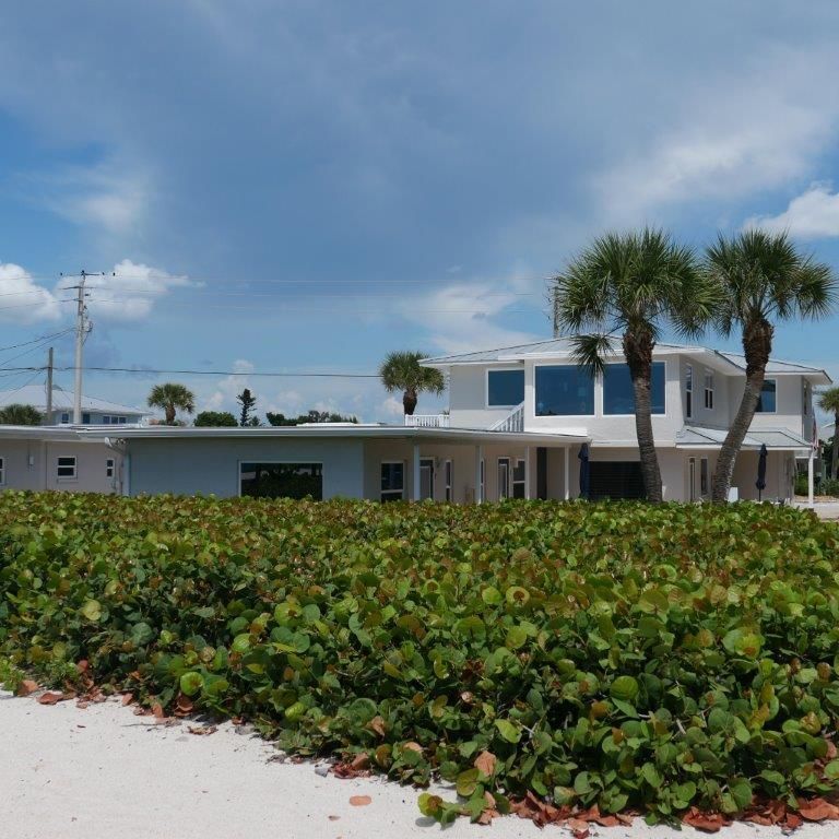Beachfront house with white sand, green shrubs, and palm trees under a partly cloudy blue sky