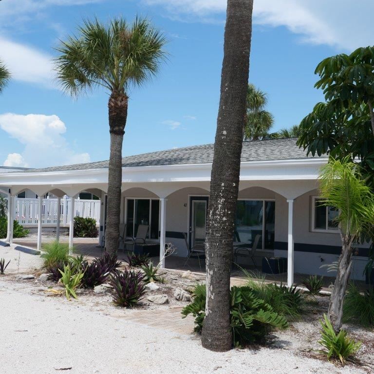 White coastal house with a shaded porch, palm trees, and sandy landscaping under a blue sky