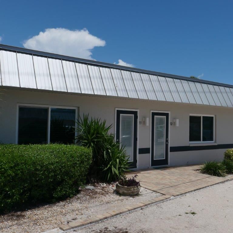 Single-story beige building with a metal roof, dark windows, and a small landscaped entrance under a blue sky